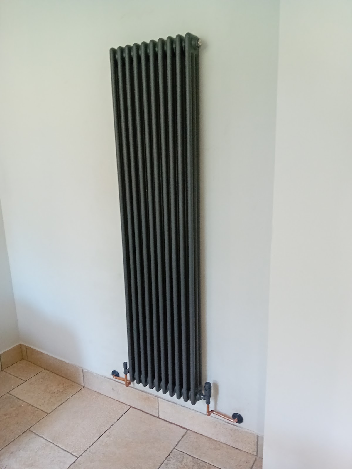 Dark anthracite vertical column radiator installed on white wall with copper pipework